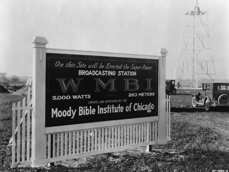 The original sign for the WMBI transmitter site stands in the forground. It says, On this site will be erected the super-power broadcasting station WMBI, owned and operated by the Moody Bible Institute of Chicago. In the background, men assemble the new tower for the transmitter.